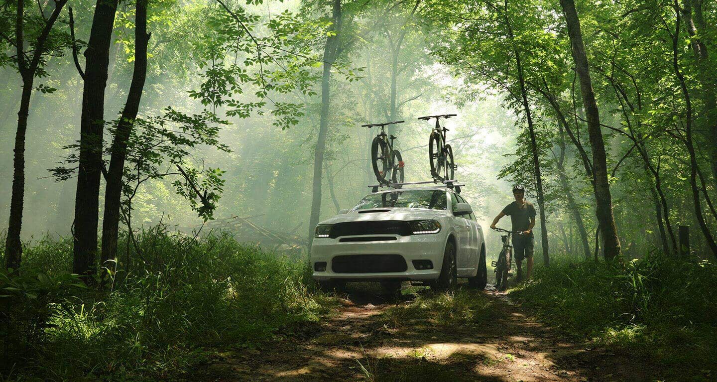 Mostrar: Vista frontal de medio perfil de un Dodge Durango 2020 estacionado en un bosque con dos bicicletas atadas a la parrilla portaequipajes.