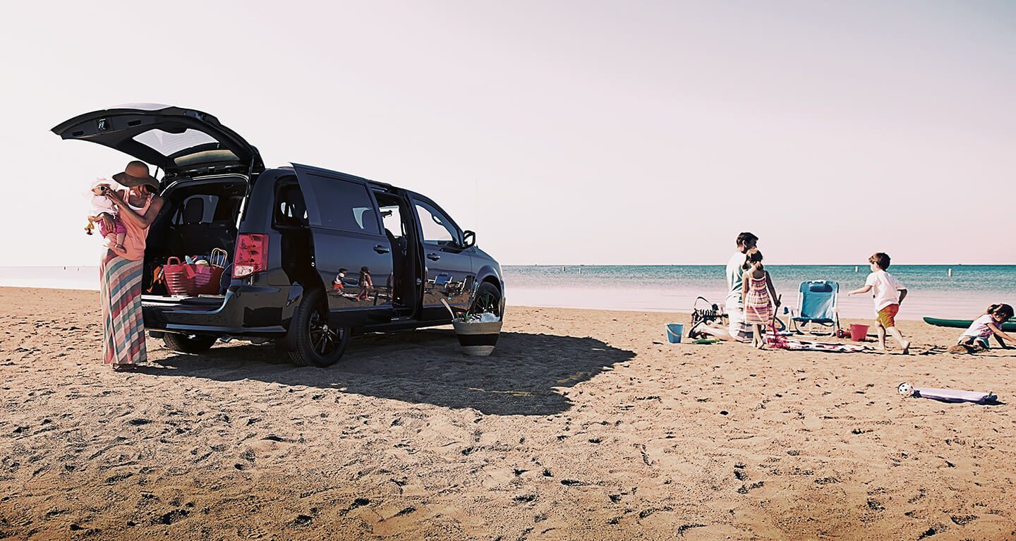 Se muestra una Dodge Grand Caravan 2020 estacionada en la playa con las puertas deslizantes y la puerta trasera abiertas y una familia descargando equipo cerca.