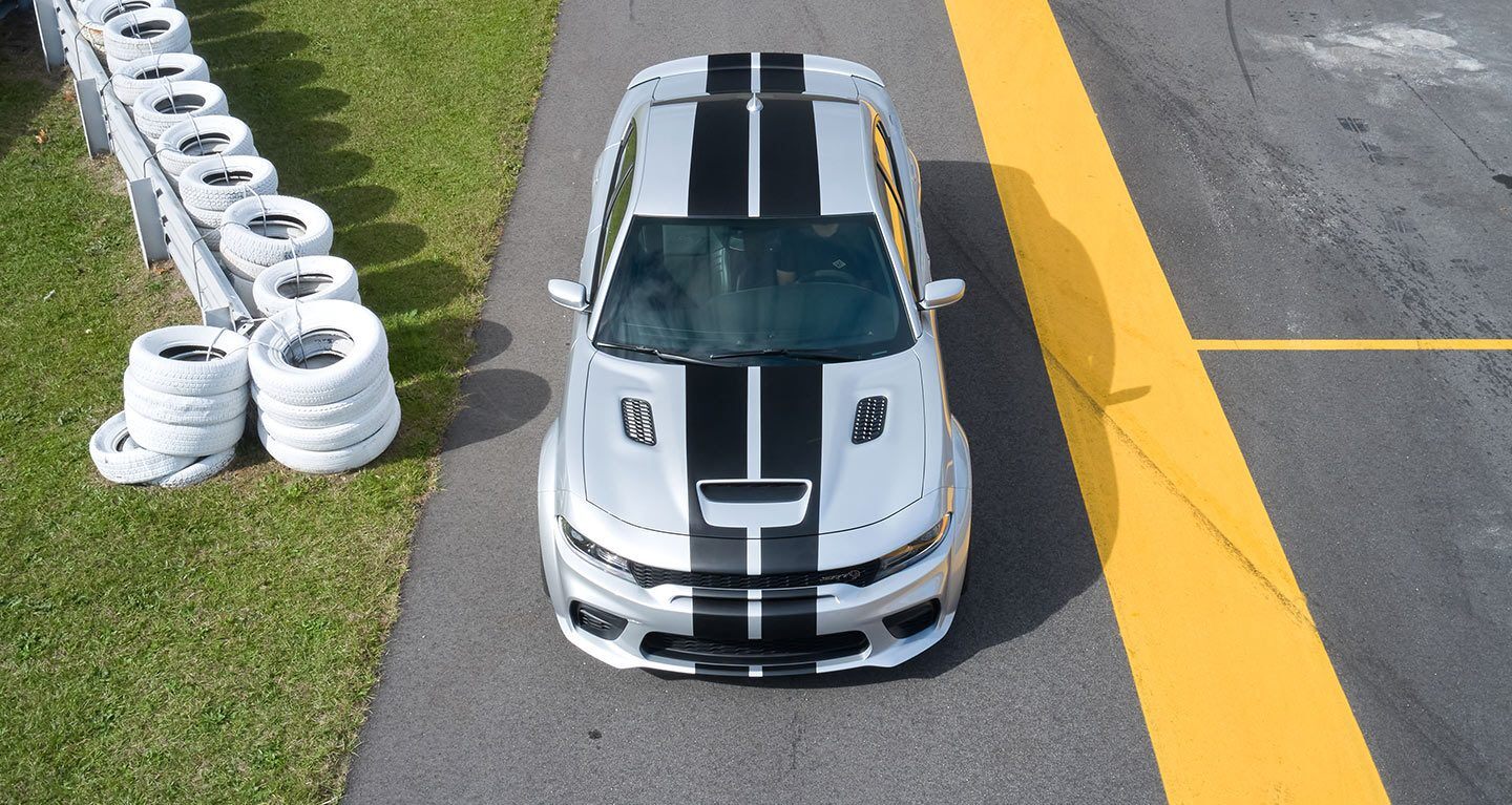 Se muestra la vista superior de un Dodge Charger SRT Hellcat Widebody 2022 con dos franjas centrales, estacionado junto a una cerca con neumáticos pintados de blanco.