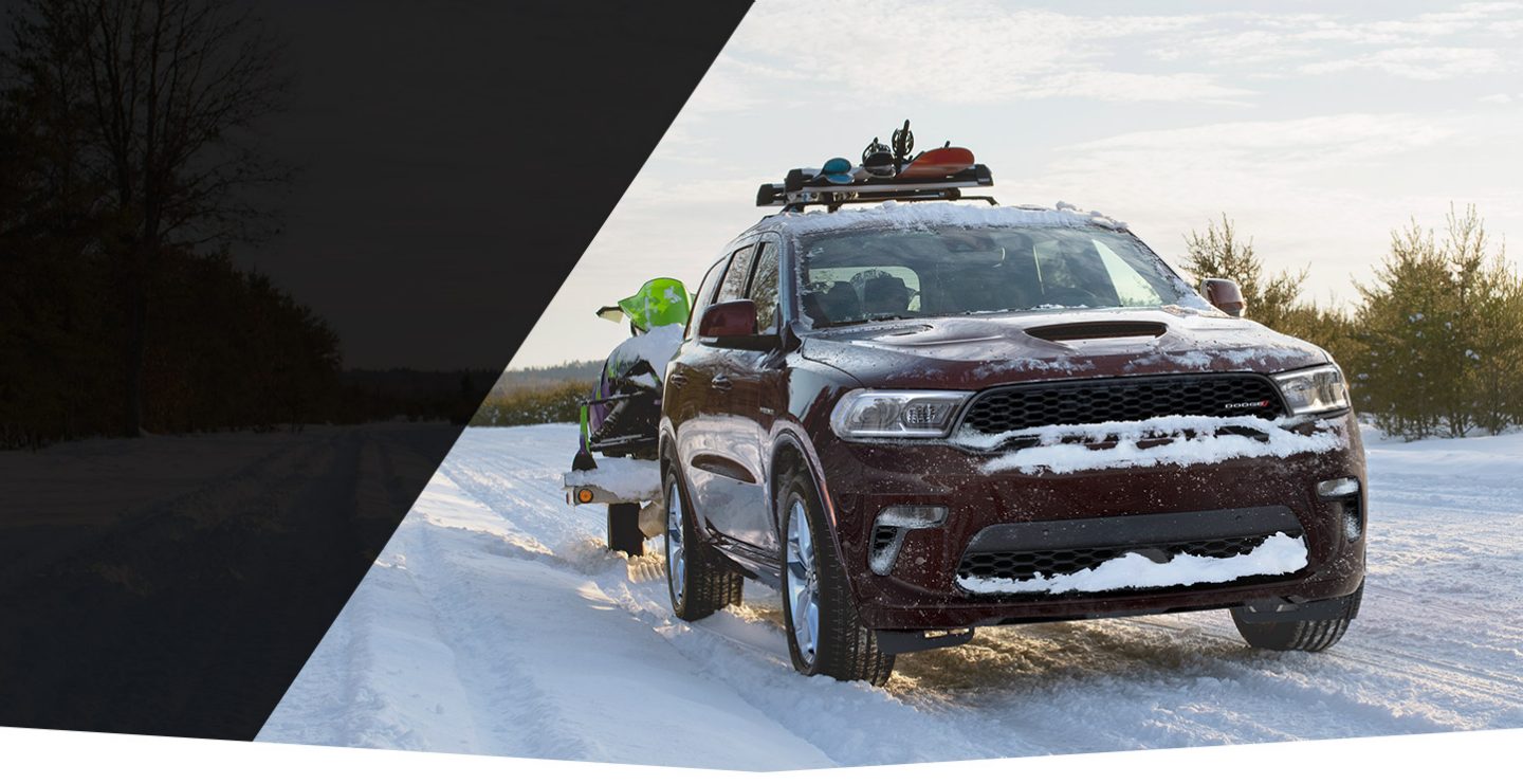 El Dodge Durango 2022 recorre un camino nevado mientras remolca un vehículo para la nieve y lleva esquíes, palos y una tabla de snowboard sobre la parrilla portaequipaje.
