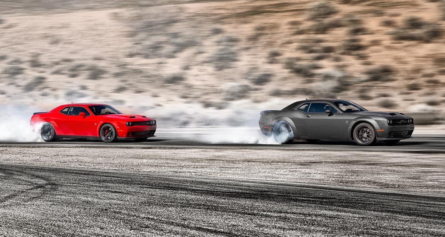 Ver Dos modelos de Dodge Challenger sobre una pista. Adelante, un Dodge Challenger SRT Hellcat 2023 gris junto a un Dodge Challenger SRT Hellcat 2023 rojo, ambos con humo saliendo de los neumáticos traseros.