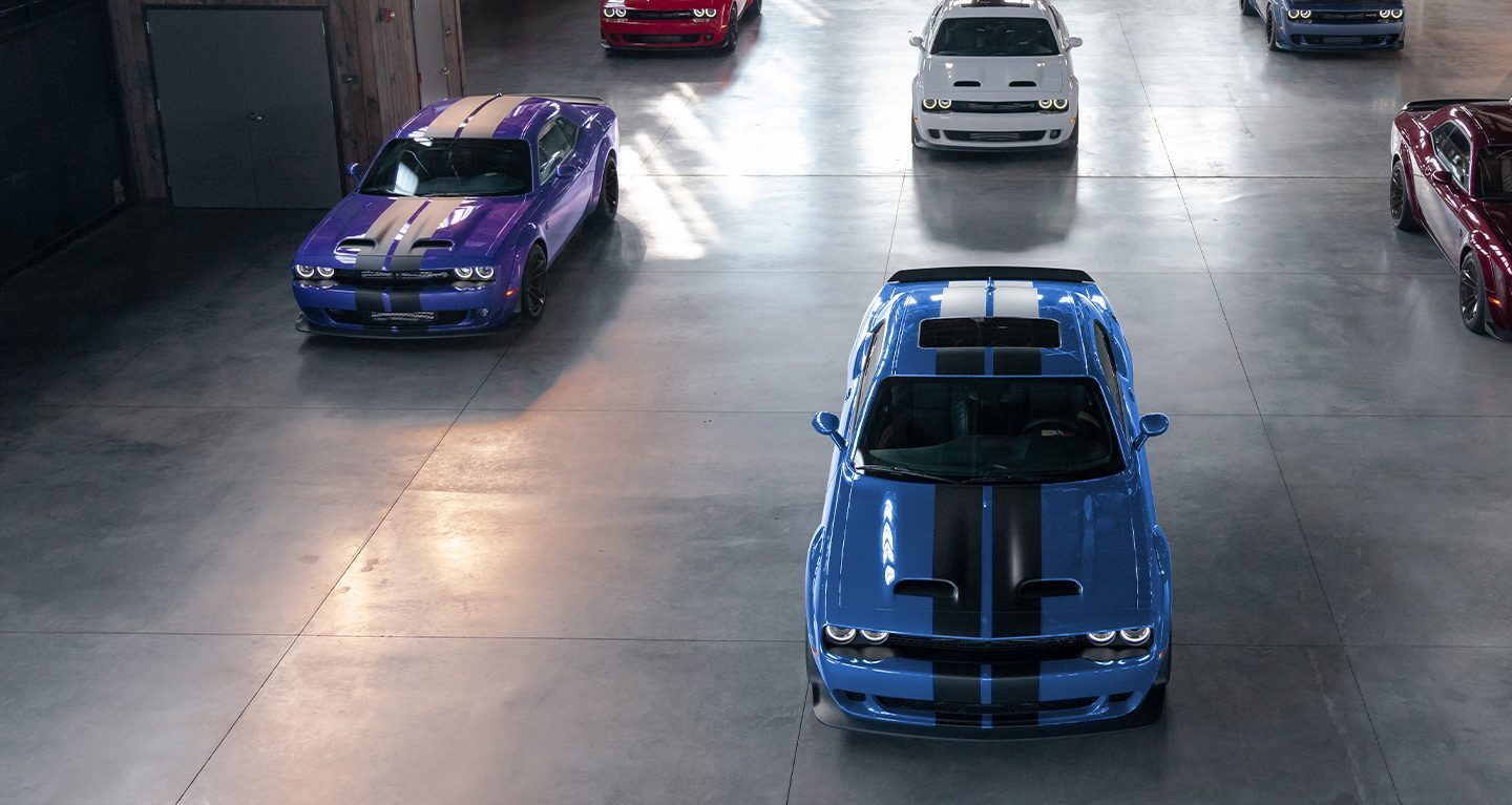 Ver Seis modelos Dodge Challenger 2023 estacionados en un garaje comercial. Entre ellos se encuentra un Challenger SRT Hellcat púrpura, un Challenger SRT Hellcat azul con rayas negras en el centro, un Challenger SRT Hellcat rojo, un Challenger SRT Hellcat blanco, etc.
