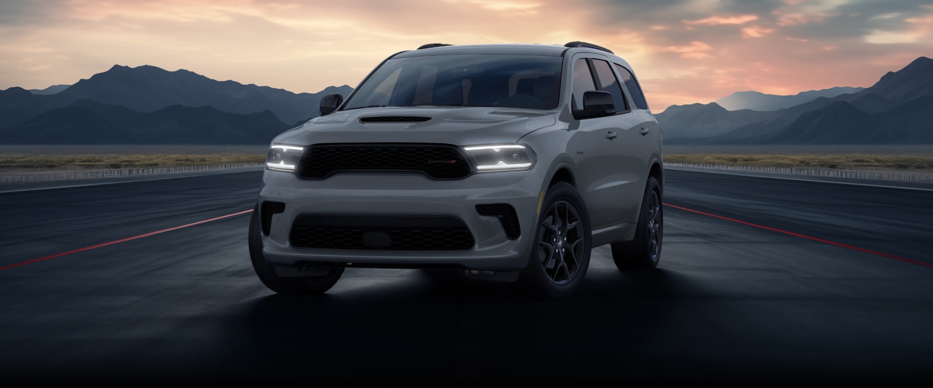 Un ángulo frontal de un Dodge Durango GT HEMI 2026 plateado estacionado en una pista rodeada de montañas al atardecer.