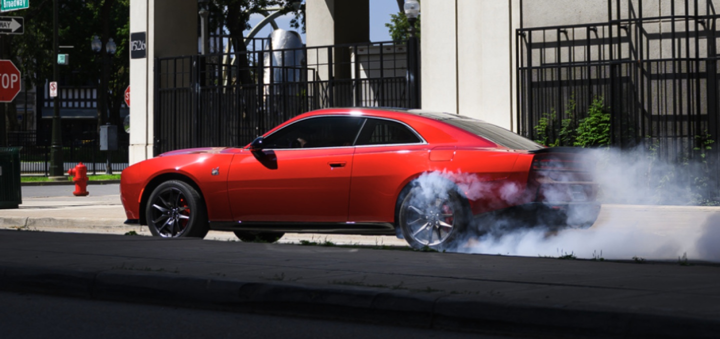 A red 2027 Dodge Charger Scat Pack in profile, stopped at an intersection, with smoke coming from its rear tires.