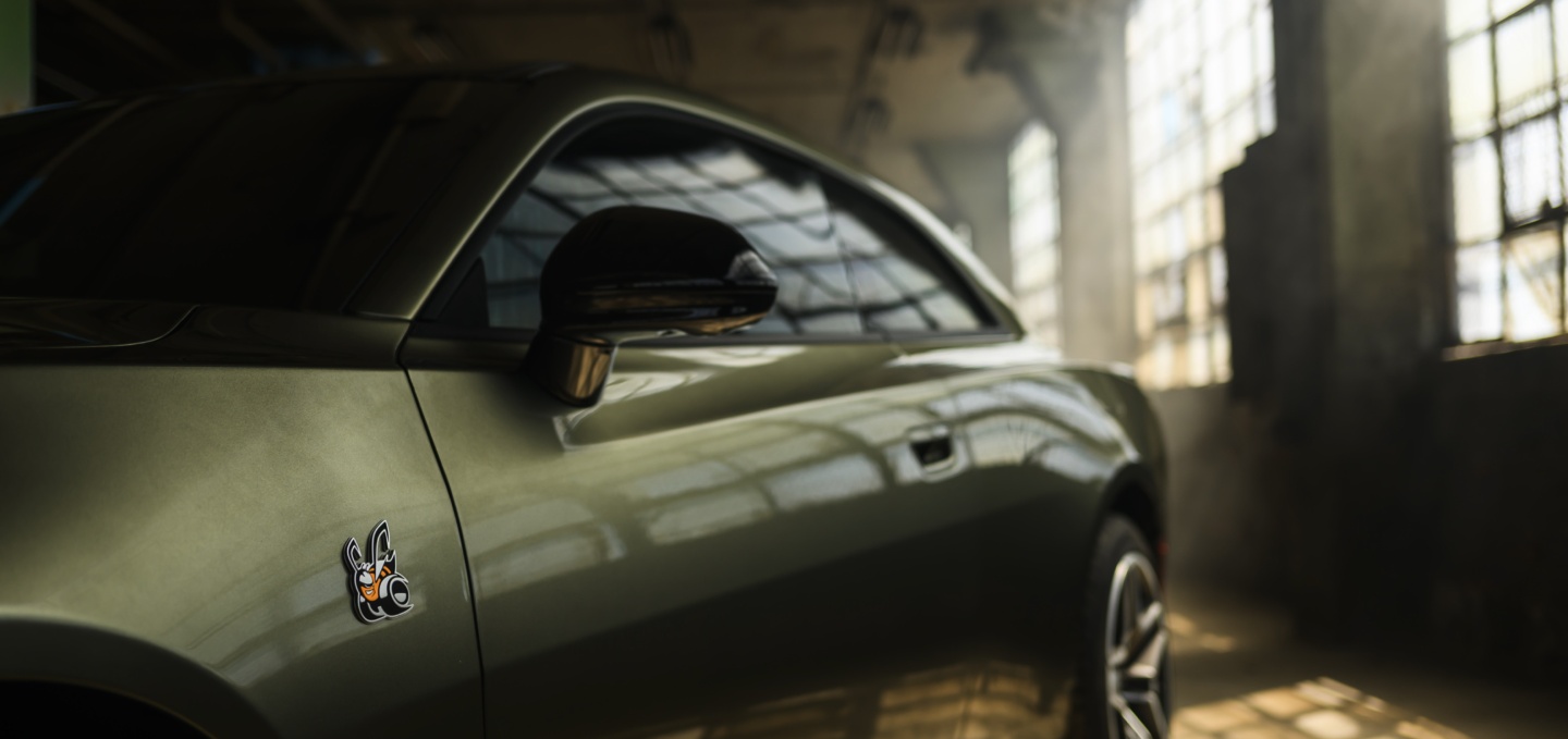 The driver's side of an olive green 2027 Dodge Charger Scat Pack, parked in an industrial garage.