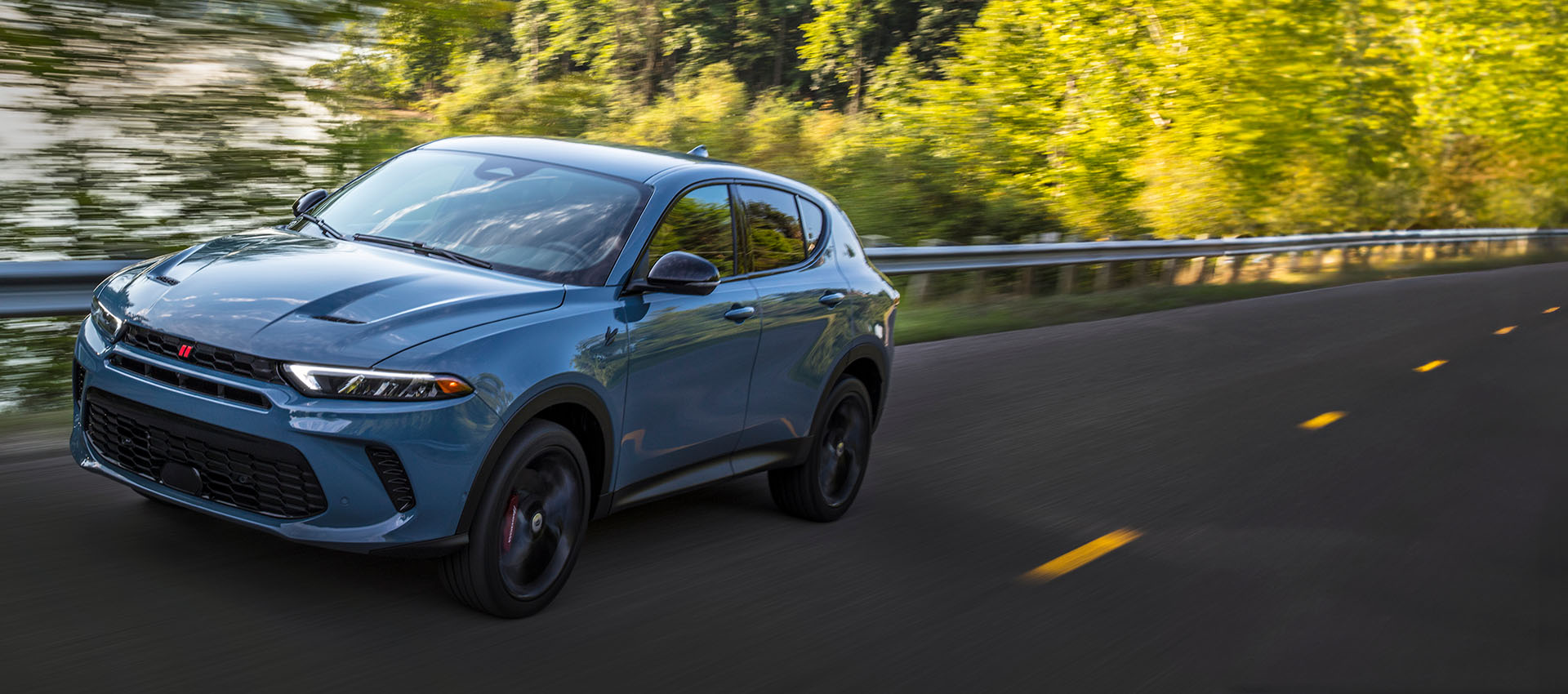 Perfil desde el ángulo del conductor de un Dodge Hornet GT 2023 azul circulando por una carretera, con el fondo borroso para indicar que el vehículo está en movimiento.