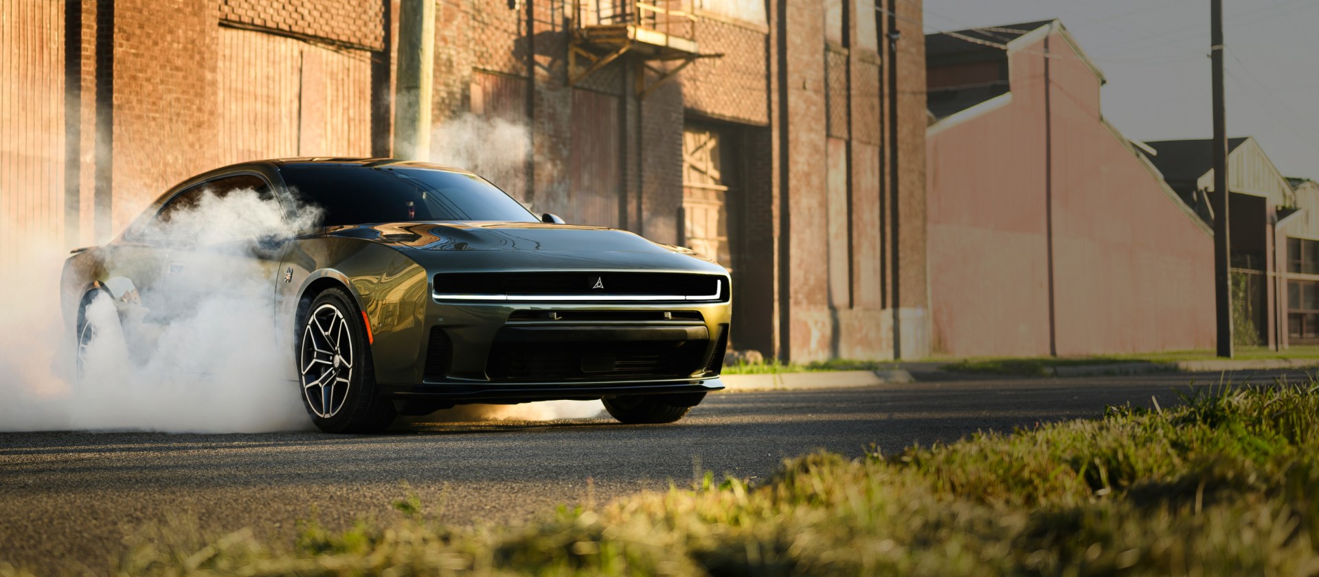 Ángulo frontal del lado del pasajero de un Dodge Charger Scat Pack Plus 2026 negro estacionado sobre asfalto con humo saliendo de los neumáticos. Al fondo se ven edificios industriales.