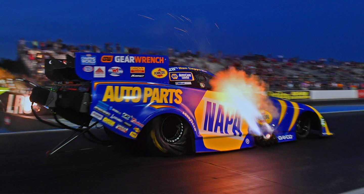 Se muestra un Funny Car azul y amarillo fotografiado al atardecer, con llamas saliendo de los tubos de escape a los costados del vehículo.