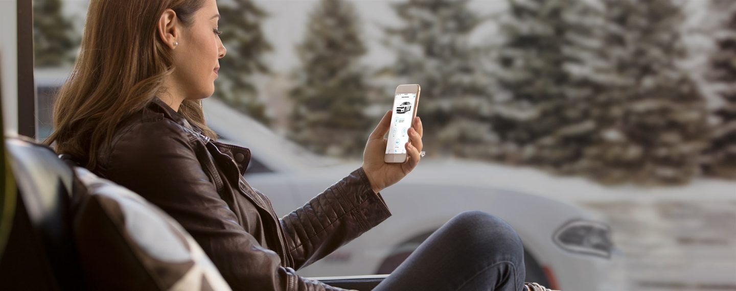Una mujer sentada al aire libre, mira la aplicación Uconnect en su smartphone, con su Dodge Charger de fondo.