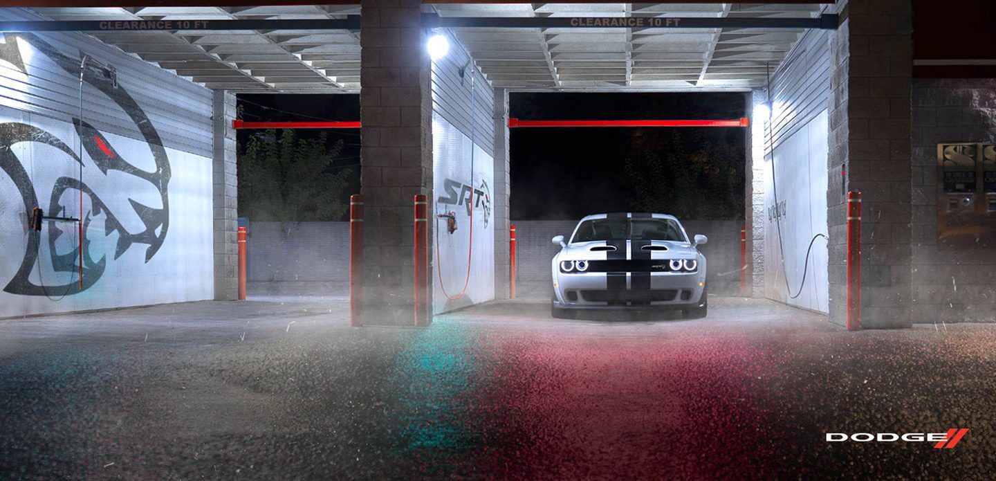 Se muestra un Dodge Challenger SRT Hellcat Redeye Widebody 2021 plateado con franjas centrales dobles en carbón, estacionado de noche en un lavadero de autos de servicio automático con gráficos SRT en las paredes.