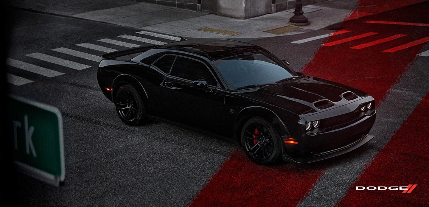 Un Dodge Challenger SRT Hellcat Widebody 2021 negro andando por una intersección, de noche, con las franjas deportivas Dodge rojas superpuestas en la calle.